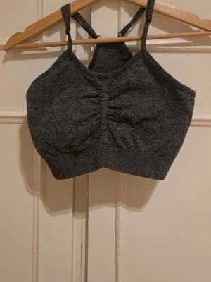Mörkgrå sport-bh med smala band - Mörkgrå sport-bh i stretchigt material med smala axelband och rynkad detalj framtill. Bh:n har en högre halsringning och bred resår under bysten för extra stöd. 
