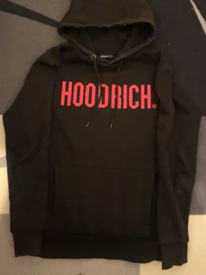 Svart HOODRICH hoodie med röd logga - Svart hoodie från Hoodrich med stor röd logotyp på bröstet. Klassisk modell med huva och dragsnören samt en stor magficka framtill. Perfekt för dig som gillar streetwear och vill sticka ut med en tydlig logga.