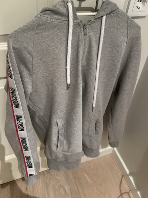 Grå hoodie från Moschino med dragkedja - Snygg grå hoodie från Moschino med dragkedja framtill och vita snören i huvan. Ärmarna har vita band med Moschino-logga och en röd linje längs sidorna. Mjuk bomullsmix och fickor framtill gör den både bekväm och stilren. Perfekt för en avslappnad streetwear-look. Har ett jätte litet hål baktill som man ser på bild 3.