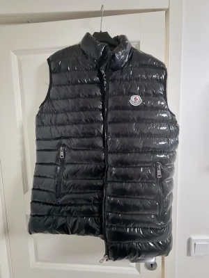 Svart dunväst från Moncler - Snygg svart dunväst från Moncler med glansig finish och klassisk logga på bröstet. Västen har två dragkedjeförsedda fickor och hög krage. Perfekt för lager-på-lager och streetwear-stil. Materialet är dunfyllning och yttertyget är blankt.
