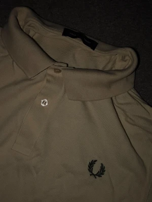 Beige pikétröja från Fred Perry - Klassisk beige pikétröja från Fred Perry med broderad logga på bröstet. Tröjan har krage, två knappar framtill och är tillverkad i ett bekvämt bomullsmaterial. Perfekt för en stilren och avslappnad look. Nypris: 1300kr
