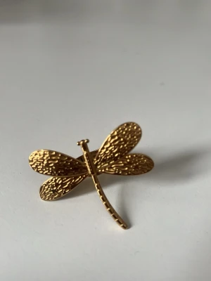 Guldfärgad trollslända ring - Unik ring i form av en trollslända i guldfärgad metall. Vingarna har en detaljerad, hamrad struktur som ger ett coolt och trendigt intryck. Perfekt accessoar för dig som gillar naturinspirerade smycken och vill sticka ut lite extra.