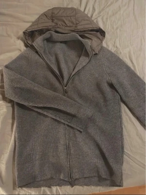 Grå stickad Cardigan med dragkedja - Snygg grå stickad Cardigan från Massimo Dutti med hel dragkedja och quiltad huva i brunt. Tröjan har ribbstickad struktur och justerbar huva med snörning. Perfekt för lager-på-lager och chill stil. Riktigt sällsynt och finns ej att köpa ny. Återförsäljs för ca 2100kr så detta är en steal.