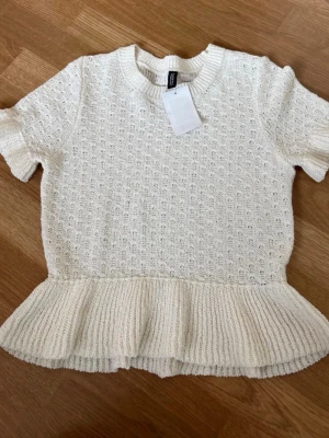 Vit stickad topp med volangkant H&M - Söt vit stickad topp från H&M med korta ärmar och volangkant nedtill. Tröjan har ett diskret hålmönster och ribbstickad rund halsringning. Perfekt för en trendig och feminin look. Materialet känns mjukt och luftigt.