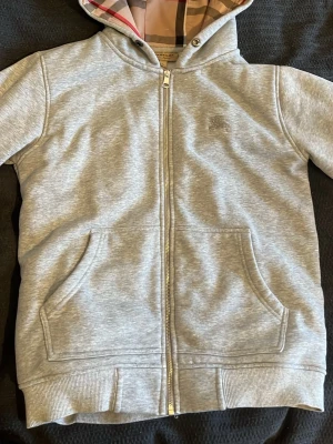 Grå zip hoodie från Burberry - Snygg grå zip hoodie från Burberry med klassiskt rutigt foder i huvan och broderad logga på bröstet. Hoodien har fickor framtill, ribbade muddar och dragkedja hela vägen. Perfekt för en chill och stilren look.