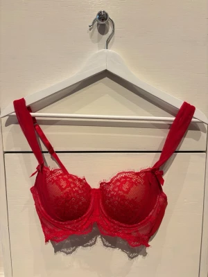 Röd spets-bh med rosetter - Snygg röd bh med spetsdetaljer och små rosetter på axelbanden från Hunkemöller. Bh:n har vadderade kupor, justerbara axelband och hakknäppning bak. Materialet är en kombination av spets och mesh som ger en feminin och trendig look. Den är aldrig använd och är som ny💕