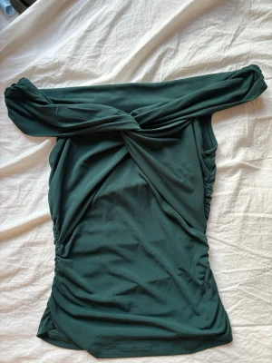 Mörkgrön off shoulder topp - Trendig mörkgrön off shoulder topp med twistad detalj framtill. Toppen har åtsittande passform och rynkade sidor som ger en snygg siluett. Tillverkad i ett stretchigt, mjukt material som känns bekvämt mot huden.