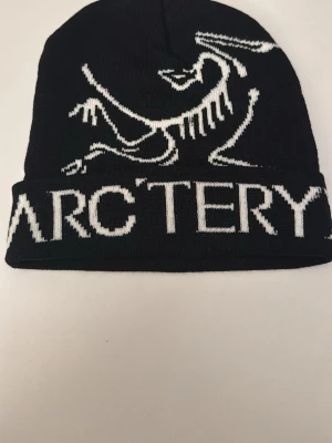 Svart mössa från Arc'teryx - Snygg svart mössa från Arc'teryx med vit logga och text runt om. Tillverkad i mjukt stickat material som sitter skönt på huvudet. Perfekt för dig som gillar streetwear och vill ha en stilren accessoar med tydlig branding.