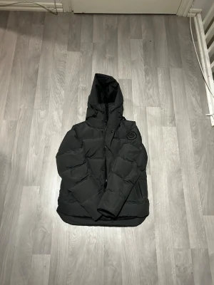 Svart pufferjacka med huva - Snygg svart pufferjacka med huva och dragkedja samt knappar framtill. Jackan har en patch med logga på bröstet och justerbara snören vid huvan. Faller fjädrar Perfekt för kalla dagar och riktigt trendig stil.