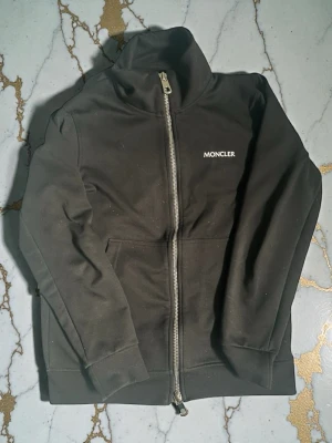 Svart zip hoodie från Moncler - Snygg svart hoodie från Moncler med dragkedja framtill och diskret Moncler-logga på bröstet. Tröjan har hög krage, långa ärmar och två fickor fram. Tillverkad i mjukt bomullsmaterial som känns skönt mot huden. Klassisk och stilren design med Moncler-märke på ärmen.