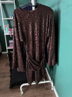 Brun glittrig kortklänning med knyt - Säljer en brun kort klänning täckt av glittriga paljetter. Klänningen har långa ärmar, rund halsringning och ett snyggt knyt i midjan som ger en cool siluett. Perfekt för dig som vill sticka ut på festen!