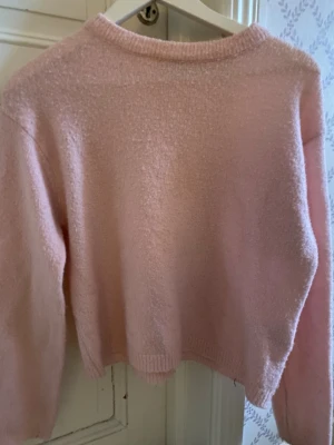 Ljusrosa stickad croppad tröja - Mysig ljusrosa stickad tröja med croppad passform och rund hals. Perfekt att styla med höga jeans eller kjol. Tröjan har långa ärmar och ribbade muddar vid hals, ärmslut och nederkant.