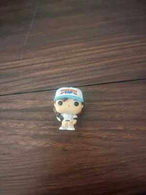 Funko Pop-figur Stranger Things - Säljer en Funko Pop-figur föreställande en karaktär från Stranger Things. Figuren har blå keps med text, vit jacka och beige skor. Perfekt för samlare eller som dekoration i hyllan.