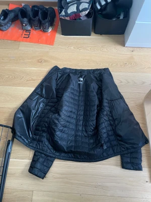Svart vindjacka från The North Face - Säljer en svart vindjacka från The North Face med quiltad design och diskret logga på bröstet. Jackan är lätt, har dragkedja och är perfekt för blåsiga dagar. Materialet är tunt och smidigt, vilket gör den enkel att packa ner i väskan.