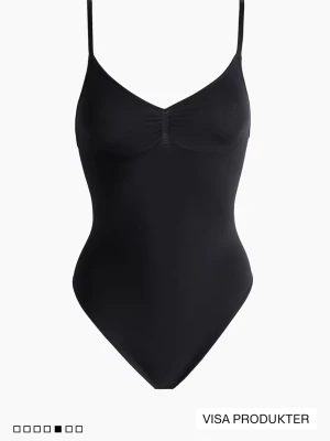 Svart sömlös body med smala band - Stilren svart body med smala, justerbara axelband och lätt rynkning vid bysten. Tillverkad i ett stretchigt, sömlöst material som ger en tight och bekväm passform. Perfekt att bära under kläder eller som ett statement-plagg.