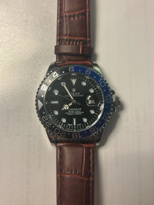 klocka med brun läderrem - Snygg klocka med svart urtavla, blå och svart vridring samt bruna läderband med krokodilstruktur. Urtavlan har tydliga markeringar och datumvisning. Klassisk och exklusiv design som sticker ut.