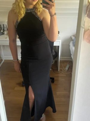 Svart långklänning med paljettdetaljer - Säljer en svart långklänning med hög hals och glittriga paljettdetaljer vid axlar och halsringning. Klänningen har en snygg slits framtill och är ärmlös, vilket ger en elegant och modern look. Perfekt för bal eller festliga tillfällen. Använt den en gång. Då jag köpte den för 700 kr är priset därför 600. 