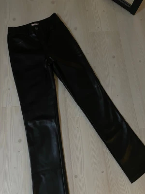 Svarta skinbyxor  - Säljer ett par svarta skinbyxor med rak passform och klassisk femficksdesign. Byxorna har medel hög midja och stängs med knapp och dragkedja. Materialet har en glansig finish som ger en cool look. Perfekta till en trendig outfit.