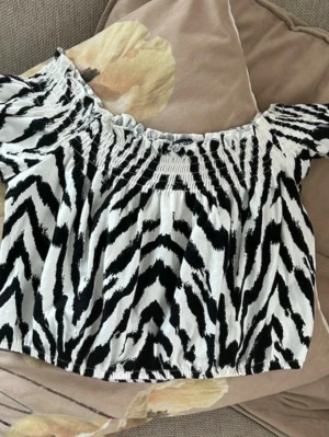 Zebramönstrad offshoulder topp Gina Tricot - Trendig offshoulder topp från Gina Tricot i svartvitt zebramönster. Toppen har smockdetaljer upptill och vid ärmslut, samt korta ärmar som sitter snyggt över axlarna. Perfekt för dig som vill sticka ut med ett djurmönster! Strl S 