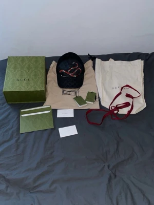 Svart Gucci keps med ormtryck - Säljer en svart keps från Gucci med GG-monogram och en stor röd orm broderad framtill. Kepsen har justerbart band bak med röd och blå rand, samt mesh på sidorna för extra ventilation. Kommer med originalbox, dustbag och tags.