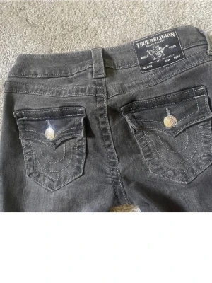 True Religion svarta low rise flare jeans - Säljer ett par svarta True Religion jeans i modellen Joey Low Rise Flare. Säljer dom då de inte kommer till användning, använda max 4 gånger. Jeansen har snygga bakfickor med dekorativa sömmar och silverfärgade knappar. Tillverkade i denim med klassisk True Religion-lapp bak i midjan. Perfekta för dig som gillar en retro vibe.