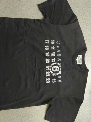 Svart t-shirt Maison Margiela - Cool svart t-shirt från Maison Margiela med det ikoniska numermotivet tryckt i vitt på bröstet. Klassisk passform och rund halsringning. Perfekt för dig som gillar streetwear och vill ha något unikt i garderoben.