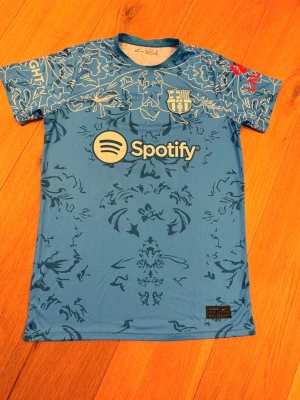 Blå FC Barcelona Nike matchtröja - Snygg blå FC Barcelona matchtröja från Nike med Spotify-logga på bröstet och klubbmärke i vitt och guld. Tröjan har ett unikt blommigt mönster över hela plagget och korta ärmar. Tillverkad i lätt och ventilerande material, perfekt för fotboll eller träning.