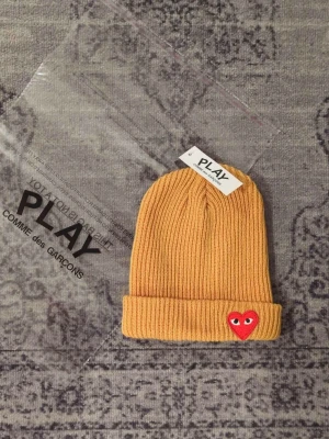 Gul ribbstickad mössa Comme des Garçons - Gul ribbstickad mössa från Comme des Garçons PLAY med uppvikt kant och det ikoniska röda hjärtat med ögon broderat framtill. Perfekt accessoar för att addera lite färg till din outfit. Klassisk och stilren design som funkar året runt.