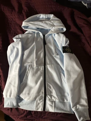 Ljusblå hoodie från Stone Island - Snygg ljusblå hoodie från Stone Island med dragkedja framtill, huva med dragsko och klassisk logotyp-patch på ärmen. Mjukt material och två fickor framtill gör den både stilren och bekväm. Perfekt för en avslappnad streetwear-look.