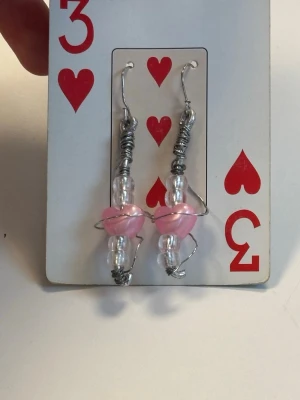 Handgjorda örhängen med rosa hjärtan - Unika örhängen med silverfärgad metalltråd, genomskinliga pärlor och rosa hjärtformade pärlor i mitten. Designen är lekfull och kreativ, perfekt för dig som gillar personliga accessoarer. Krokar i metall för enkel användning.