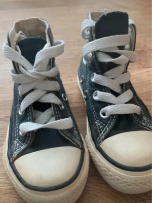 Converse All Star blåa high tops - Klassiska Converse All Star high top sneakers i blå canvas med vita gummisulor och tåhättor. Skorna har vita snören och tydliga kontrastsömmar samt den ikoniska loggan på sidan. Perfekta för en avslappnad och cool stil.