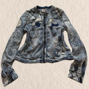Unik jeansjacka med stjärnor - Unik ljusblå jeansjacka med patchade detaljer, stjärntryck på ärmarna och rutmönster på axlar och ärmslut. Jackan har dragkedja framtill, figurnära passform och coola broderade märken. Perfekt för dig som vill sticka ut med din stil.