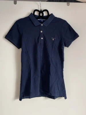 Marinblå pikétröja från Gant - Klassisk marinblå pikétröja från Gant med korta ärmar och krage. Tröjan har knappar framtill och en liten broderad logga på bröstet. Tillverkad i mjuk bomull som känns skön mot huden. Perfekt till jeans eller kjol.