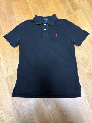 Svart pikétröja från Ralph Lauren m 10-12 år - Klassisk svart pikétröja från Ralph Lauren med röd broderad logga på bröstet. Tröjan har korta ärmar, krage och knappar framtill. Perfekt för en stilren och avslappnad look.