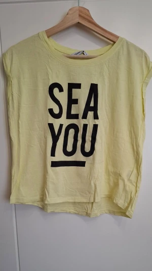 Gul t-shirt med tryck från Cropp - Gul t-shirt från Cropp med svart texttryck 'SEA YOU' på framsidan. T-shirten har rund halsringning och ärmlös design, vilket ger en avslappnad vibe. Tillverkad i mjuk bomull som känns skön mot huden. Perfekt för dig som gillar statement-plagg.