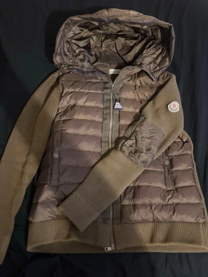 moncler cardigan - Säljer en olivgrön pufferjacka från Moncler med stickade ärmar och huva. Jackan har dragkedja framtill, två fickor med tryckknappar och en Moncler-logga på ärmen. Materialmix med quiltad polyester och stickade detaljer ger en modern look. NFC SCAN