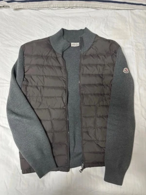 Grå cardiganjacka från Moncler - Snygg grå cardiganjacka från Moncler med quiltad front och stickade ärmar. Jackan har dragkedja framtill och en klassisk Moncler-logga på ena ärmen. Perfekt för lager-på-lager och riktigt stilren look. Storlek M