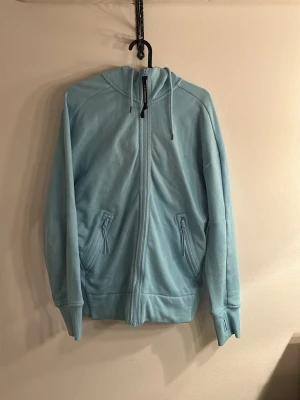 Ljusblå hoodie från C.P. Company - Säljer en ljusblå hoodie från C.P. Company med dragkedja framtill och två fickor med dragkedja. Huvan har inbyggda glasögon, vilket ger en unik och cool look. Tillverkad i mjukt bomullsmaterial och har ribbade muddar vid ärmslut och nederkant.