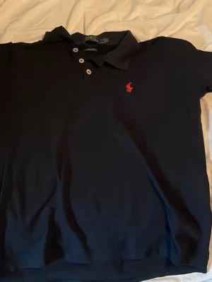  Ralph Lauren Piké - Snygg marinblå pikétröja från Ralph Lauren med röd broderad logga på bröstet. Tröjan har klassisk krage, tre knappar och är i en slim fit-modell. Perfekt för dig som gillar stilrena och tidlösa plagg. Den är i M men skulle säga att den passar S eller till och med Xs. 