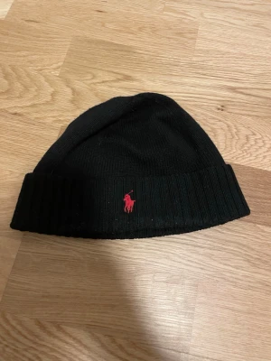 Svart mössa från Polo Ralph Lauren - Svart ribbstickad mössa från Polo Ralph Lauren med uppvikt kant och röd broderad logga framtill. Klassisk och enkel design som passar till det mesta. Tillverkad i mjukt material som håller dig varm under kalla dagar.