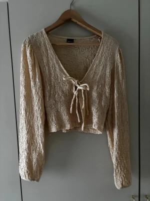 Beige omlottopp från Gina Tricot - Trendig beige omlottopp från Gina Tricot med lång ärm och knyt framtill. Toppen har en croppad passform och är gjord i ett strukturerat, lätt material som ger en cool look. Perfekt att styla med höga jeans eller kjol.