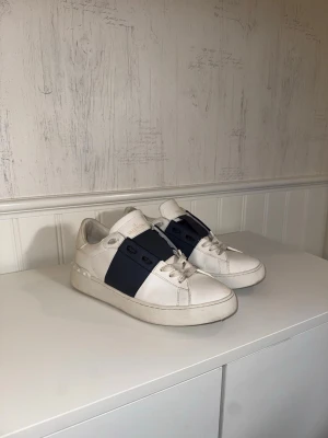 Valentino Garavani vita sneakers - Säljer ett par vita Valentino Garavani sneakers med marinblå detalj över snörningen. Använda sparsamt en sommar. Går säkert att rengöra mer noggrant om man har rätt medel! 