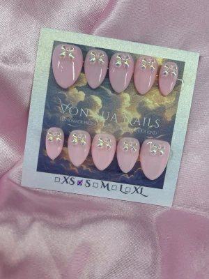 BabyBow (S);Short Almond - Snygga handgjorda press-on naglar i en mjuk rosa nyans. Naglarna har en kort almondform och är dekorerade med holo-rosetter för en extra söt touch. Perfekta för dig som vill ha ett gulligt och trendigt nagelset. S: 15 mm, 11,5 mm, 12 mm, 11 mm, 9 mm 