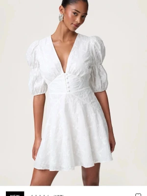 Vit kort klänning med puffärmar - Säljer en vit kort klänning med v-ringning, puffiga kortärmar och dekorativa knappar framtill. Klänningen har ett subtilt blommigt mönster och markerad midja. Perfekt för sommarens alla tillfällen! Aldrig använd prislapp sitter kvar. 