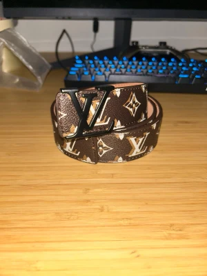 Brunt Luis Viton monogram skärp - Snyggt brunt skärp från Lois Vuton med klassiskt monogrammönster och stor L&V-spänne i svart metall. Skärpet har en exklusiv känsla och är tillverkat i präglat skinn med vita och beige detaljer i mönstret.