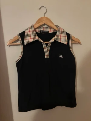 Burberry ärmlös topp med rutig krage - Snygg svart ärmlös topp från Burberry med klassisk beige-rutig krage och detaljer vid ärmöppningarna. Toppen har en liten Burberry-logga broderad på bröstet och en v-ringad öppning framtill.