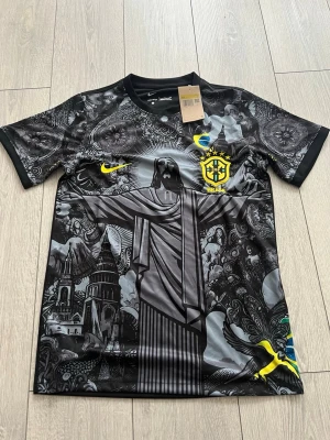 Nike Brasil svart grafisk fotbollströja - Snygg svart fotbollströja från Nike med Brasilien-tema. Tröjan har ett stort grafiskt tryck med Kristusstatyn, kyrkor och detaljerade mönster. Brasilienmärket och Nike-loggan i gult på bröstet. Tillverkad i lätt, andningsbart material perfekt för träning eller match.