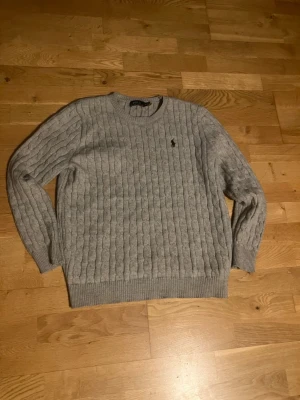 Grå kabelstickad tröja från Polo Ralph Lauren - Kabelstickad grå tröja från Polo Ralph Lauren med rund halsringning och diskret broderad logga på bröstet. Tröjan har ribbade muddar vid ärmslut och nederkant, och är tillverkad i mjukt material som passar perfekt till höst och vinter.