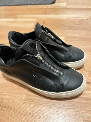 Svarta Arigato sneakers med dragkedja - Snygga svarta sneakers från Arigato i läder med unik dragkedja framtill istället för klassiska snören. Vit sula med tydlig logga undertill. Minimalistisk design och diskret branding på sidan. Perfekta för dig som gillar stilrena och moderna skor.