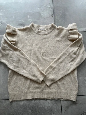 Beige stickad tröja med volangärm - Beige stickad tröja från Unistudio med rund halsringning och långa ärmar. Tröjan har unika detaljer med volanger på axlarna och ribbstickade muddar vid ärmslut och nederkant. Perfekt för dig som gillar en enkel men ändå trendig look. Bara andvänd nån gång.